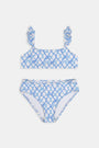 A young girl with curly hair smiles in the Roller Rabbit Kids Seaside Knots Rory Bikini, featuring ruffled shoulder straps and UPF 50+ sun protection, posing against a plain light background.