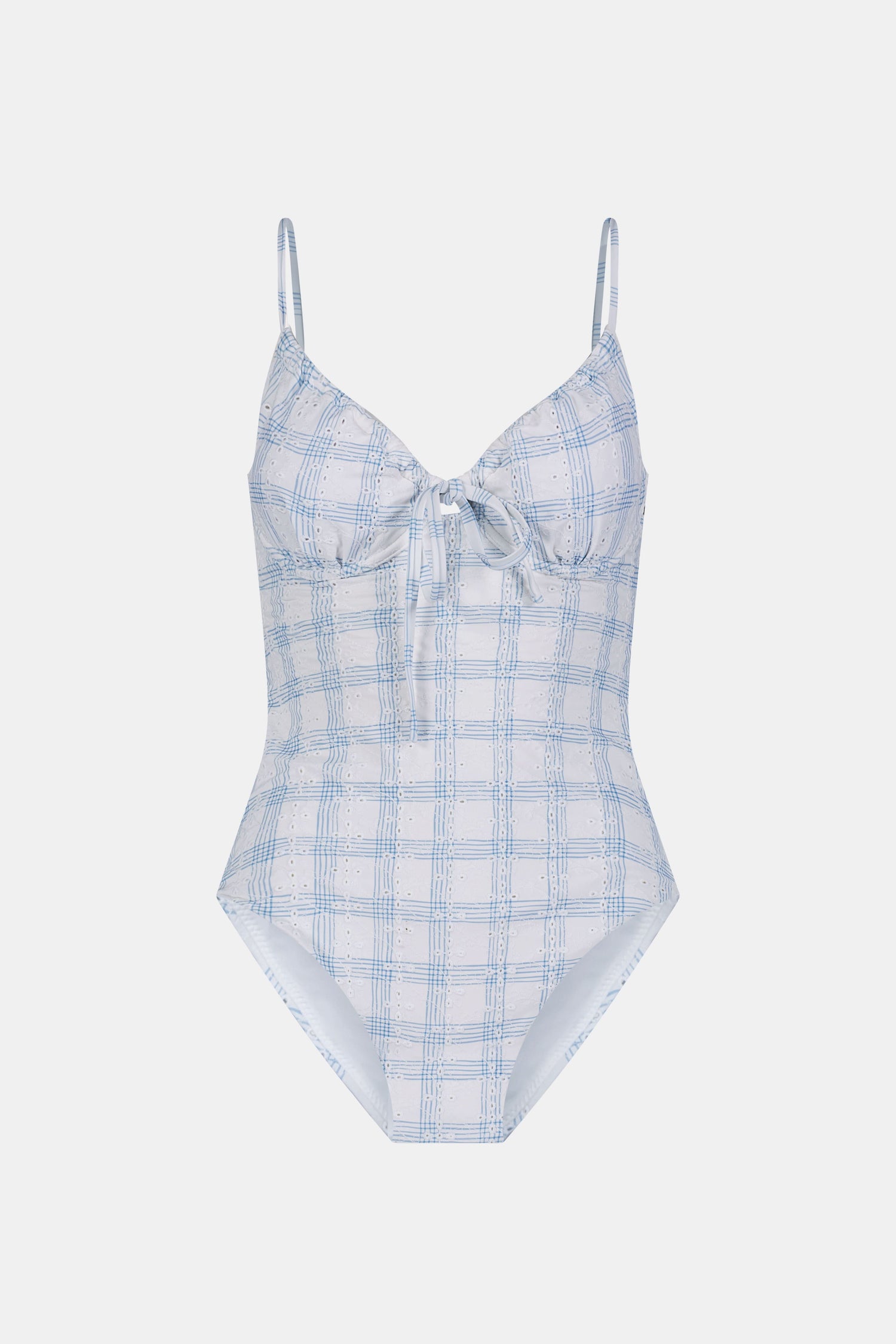 A woman wearing the Roller Rabbit Sun Check Eyelet Aurora One Piece, a light blue and white plaid swimsuit with adjustable straps, stands on a beach with waves and a blue sky in the background.
