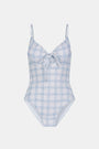 A woman wearing the Roller Rabbit Sun Check Eyelet Aurora One Piece, a light blue and white plaid swimsuit with adjustable straps, stands on a beach with waves and a blue sky in the background.