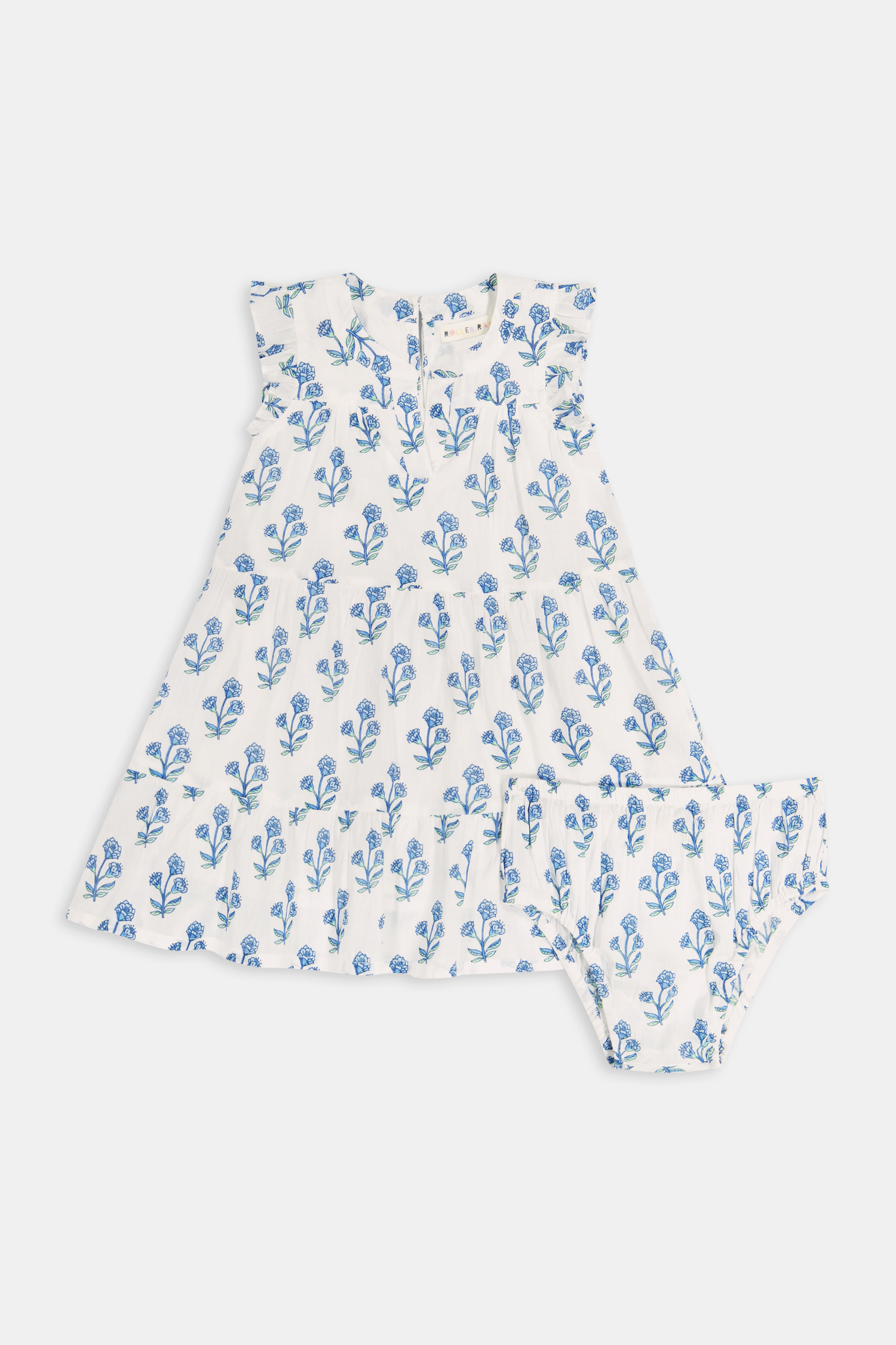 A young girl with long brown hair wears the Roller Rabbit Kids Rosa Ditsy Floral Pami Dress, made from cotton voile with blue-green block print and ruffled sleeves, standing against a plain light background, looking slightly to the side.