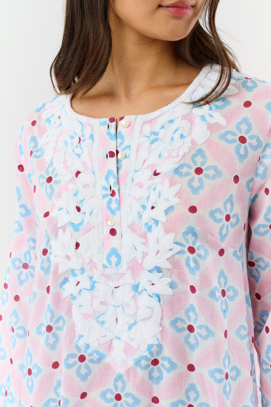 A woman with long hair wears the Roller Rabbit Lerato Inez Long Kurta, a cotton voile long sleeve pullover dress featuring a pastel pink and blue geometric pattern and white embroidery on the chest. She pairs it with white open-toe sandals.