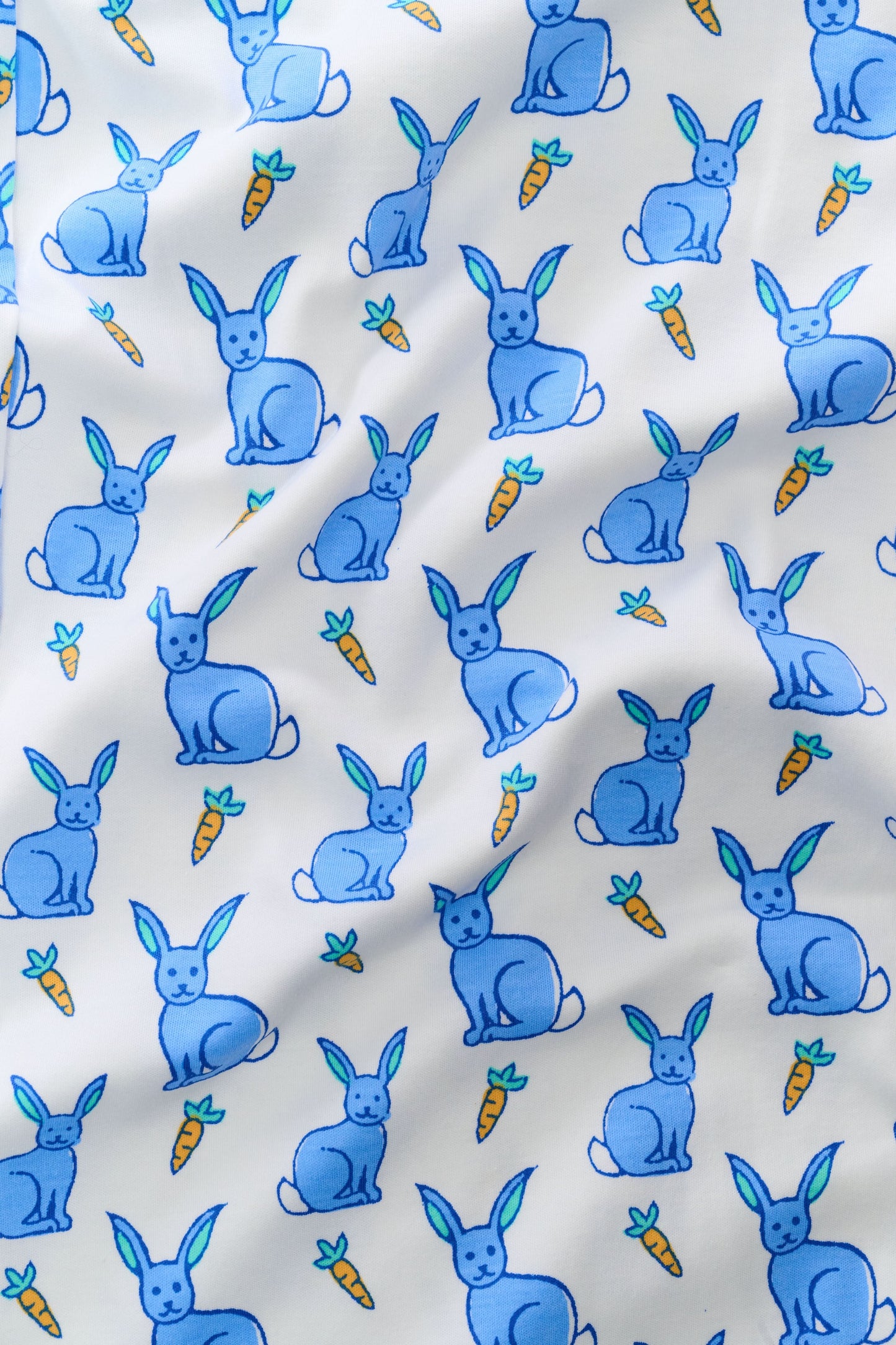 A woman with long brown hair stands barefoot in the Roller Rabbit Romy the Rabbit Long Sleeve Polo Pajamas—a matching white set with a blue bunny print, featuring a button-up top and loose-fitting pants. She has a neutral expression.