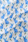 A woman with long brown hair stands barefoot in the Roller Rabbit Romy the Rabbit Long Sleeve Polo Pajamas—a matching white set with a blue bunny print, featuring a button-up top and loose-fitting pants. She has a neutral expression.