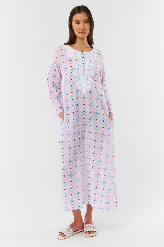 A woman with long hair wears the Roller Rabbit Lerato Inez Long Kurta, a cotton voile long sleeve pullover dress featuring a pastel pink and blue geometric pattern and white embroidery on the chest. She pairs it with white open-toe sandals.