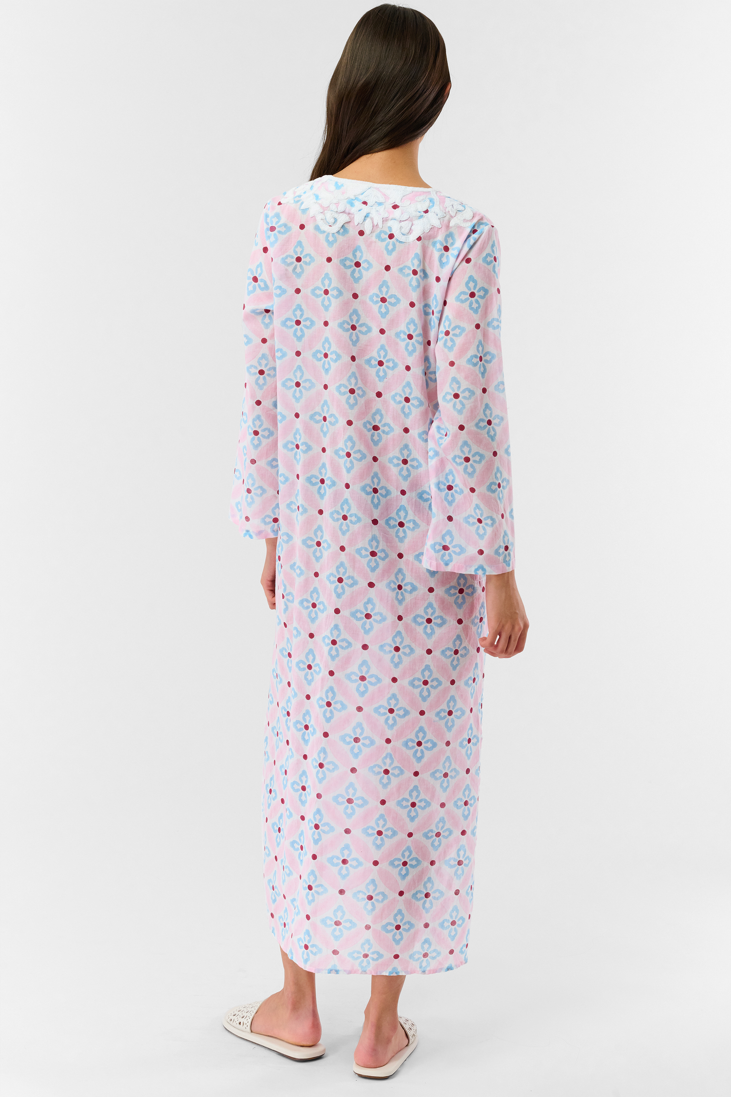 A woman with long hair wears the Roller Rabbit Lerato Inez Long Kurta, a cotton voile long sleeve pullover dress featuring a pastel pink and blue geometric pattern and white embroidery on the chest. She pairs it with white open-toe sandals.