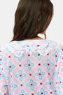 A woman with long hair wears the Roller Rabbit Lerato Inez Long Kurta, a cotton voile long sleeve pullover dress featuring a pastel pink and blue geometric pattern and white embroidery on the chest. She pairs it with white open-toe sandals.