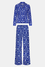 A man stands barefoot in Roller Rabbit’s Mens Lobster Trellis Loungewear, a matching blue and white floral cotton voile pajama set with a button-up long-sleeve shirt and pants. He has one hand in his pocket and looks forward.