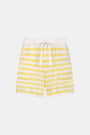 A person wears Roller Rabbit’s Striped Brie Short—a knit yellow and white horizontal striped short with a white drawstring at the elastic waistband and hands tucked in the front pockets.