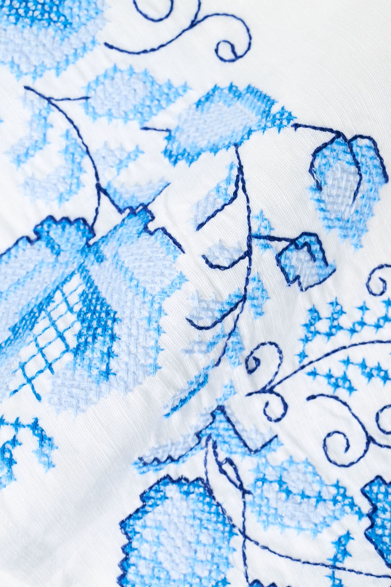 A woman wears the Roller Rabbit Cross Stitch Vine Allora Dress, a short white linen dress with blue floral embroidery on the sleeves and hem, standing with one hand in her pocket against a plain background.