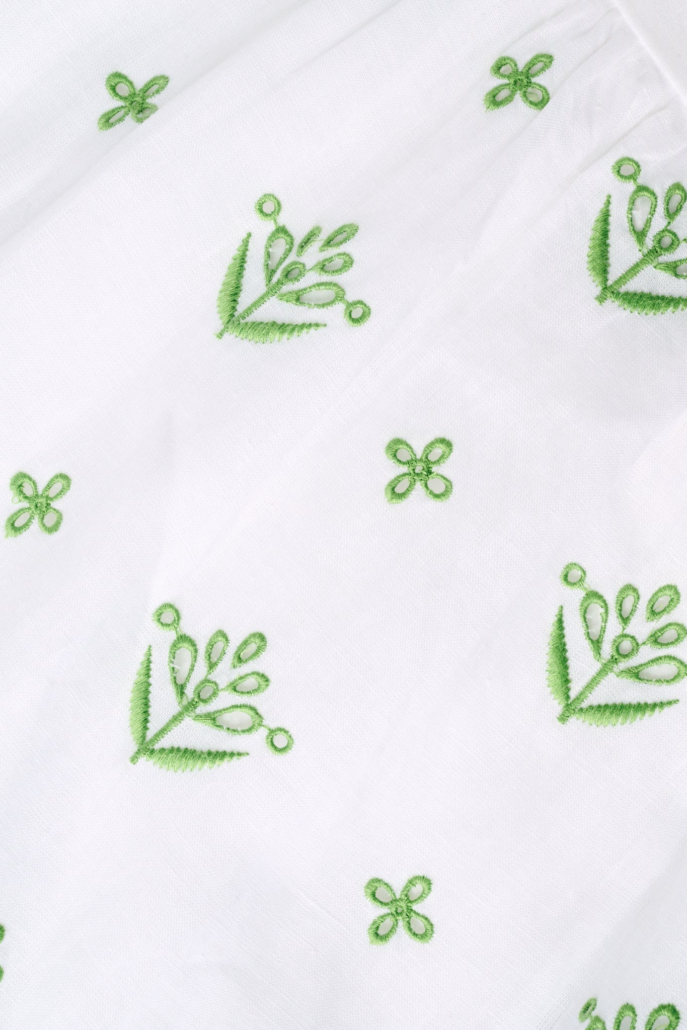 A woman with curly hair wears the Roller Rabbit Fruit Eyelet Selena Dress, a green linen maxi with floral embroidery and thin straps. She stands facing forward in yellow open-toe sandals against a plain white background.