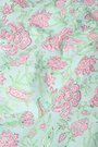 A woman with curly hair wears the Camellia Block Keaton Dress by Roller Rabbit, a sleeveless, loose-fit 100 cotton sundress with pink floral print on a light green background. She smiles softly with her hands behind her back.