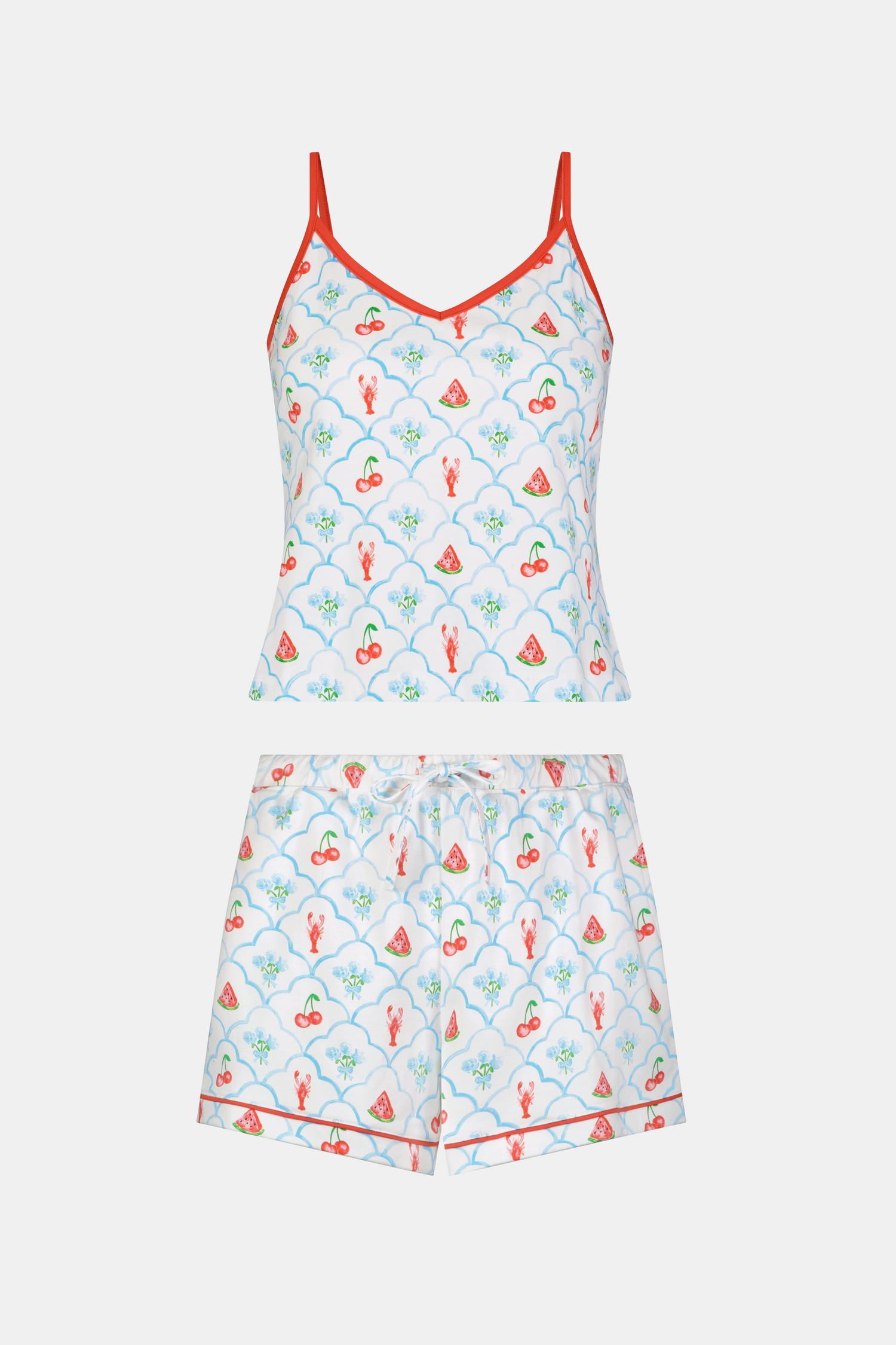 A woman with curly hair wears the Roller Rabbit 4th of Jemina Mischa Set, a Pima Cotton cami and shorts loungewear set with a white base, colorful fruit and flower prints, red trim, and drawstring shorts edged in red.