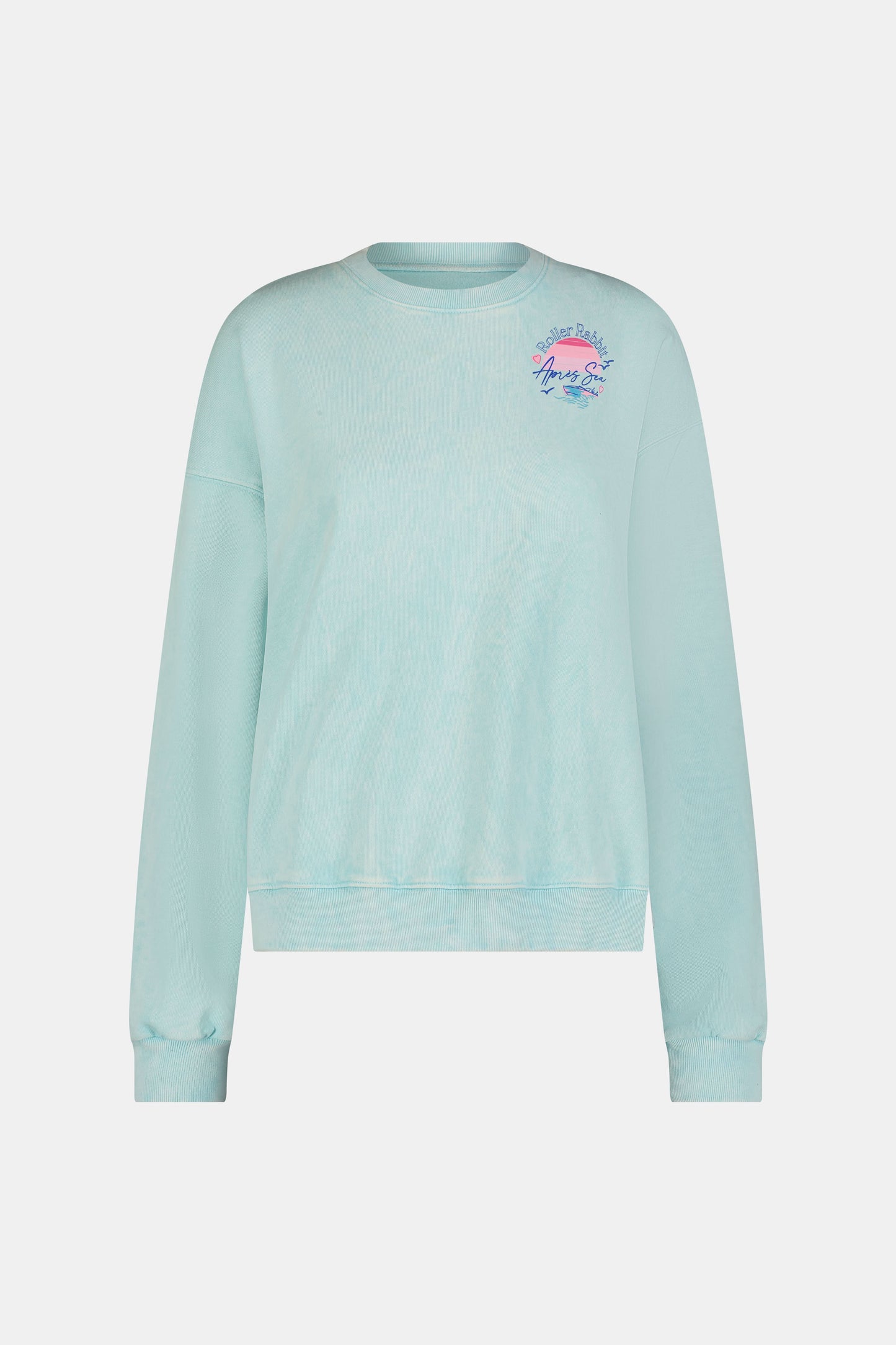 A woman wears the Roller Rabbit Apres Sea Phoebe Sweatshirt, made in the USA, mint green with a pink sunset, blue waves, birds, and "Roller Rabbit Après Sea" text on the upper left chest.