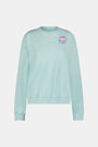A woman wears the Roller Rabbit Apres Sea Phoebe Sweatshirt, made in the USA, mint green with a pink sunset, blue waves, birds, and 