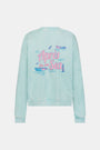 A woman wears the Roller Rabbit Apres Sea Phoebe Sweatshirt, made in the USA, mint green with a pink sunset, blue waves, birds, and 