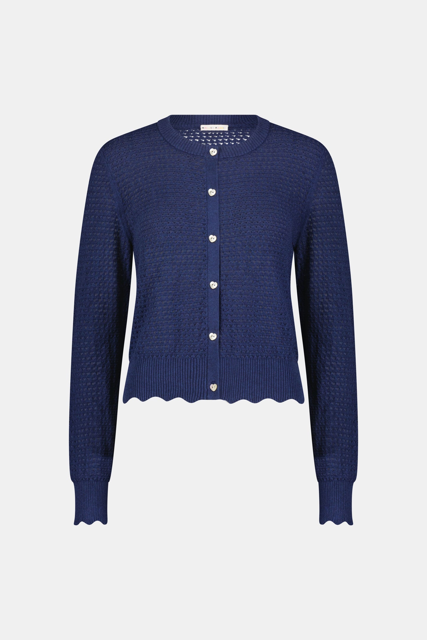 A woman wears the Roller Rabbit Pointelle Rosella Cardigan, a navy blue button-up with heart-shaped buttons, paired with white pants featuring a blue floral print, standing against a plain light background.