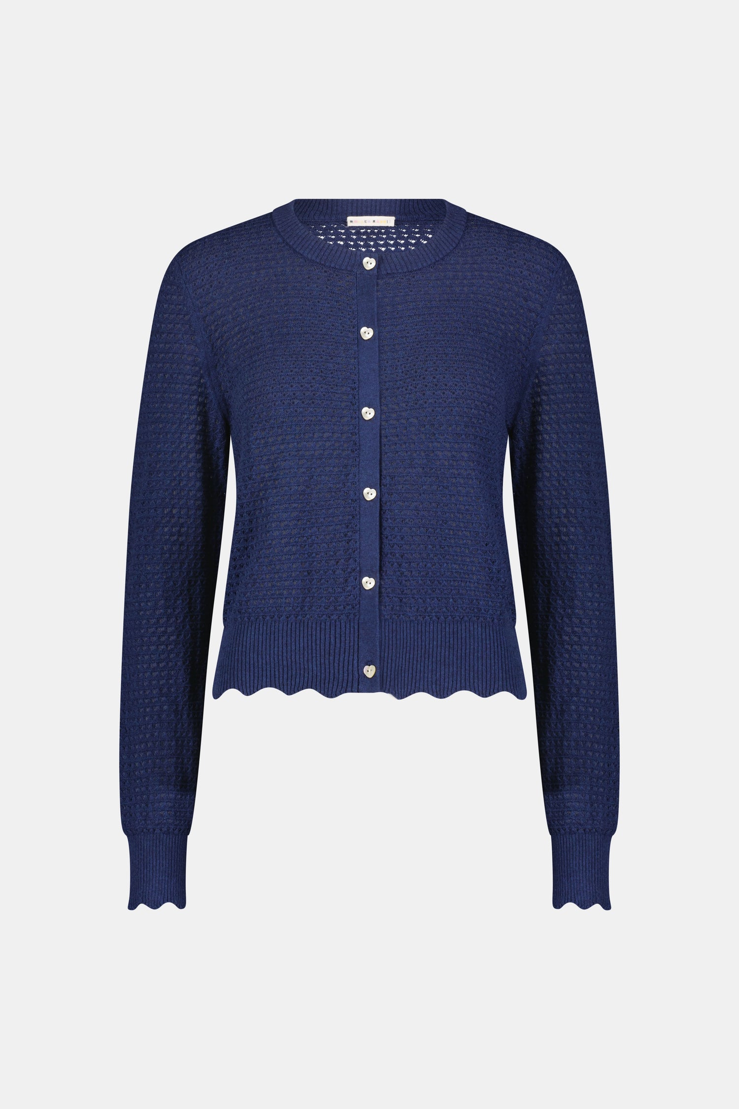 A woman wears the Roller Rabbit Pointelle Rosella Cardigan, a navy blue button-up with heart-shaped buttons, paired with white pants featuring a blue floral print, standing against a plain light background.