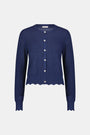 A woman wears the Roller Rabbit Pointelle Rosella Cardigan, a navy blue button-up with heart-shaped buttons, paired with white pants featuring a blue floral print, standing against a plain light background.