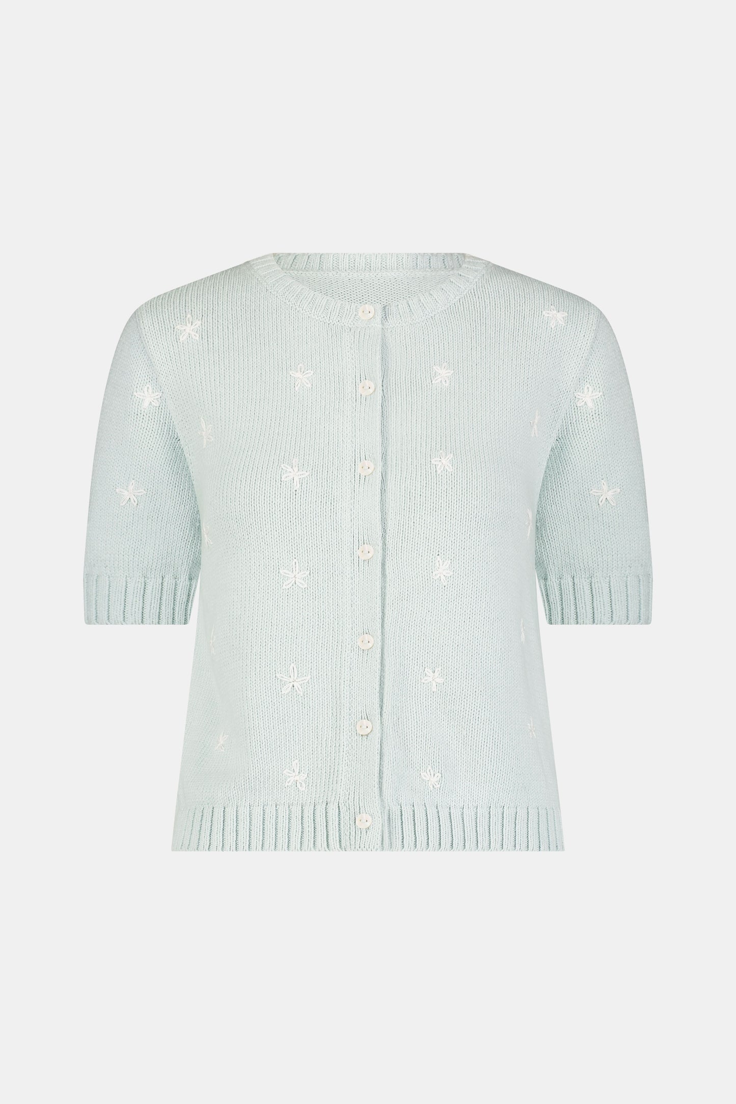 Close-up of a woman wearing the Roller Rabbit Floral Bloom Nyla Cardigan, a light blue cotton button-up with white embroidered stars, rounded neckline, and short sleeves, styled with white eyelet shorts.