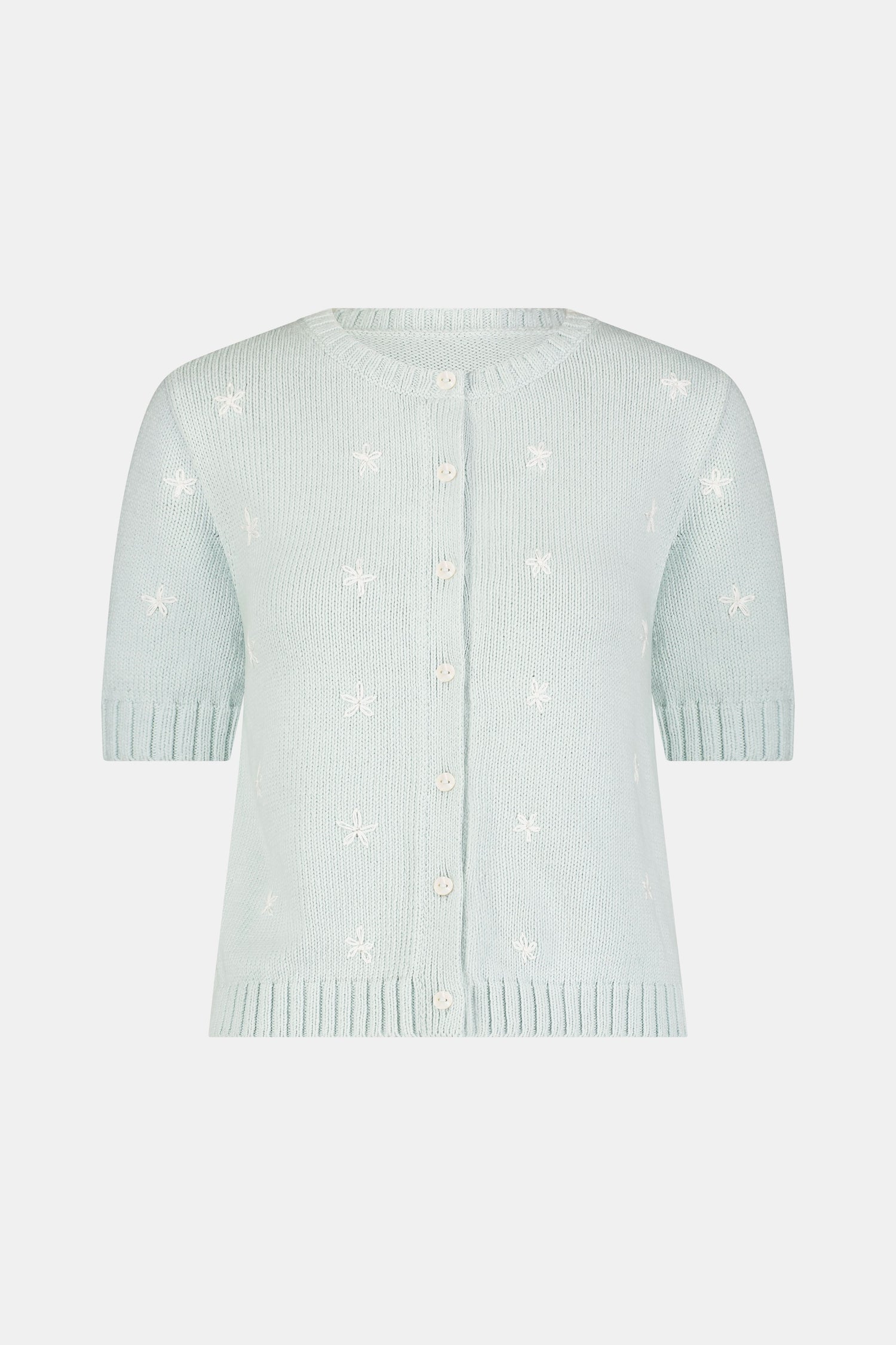Close-up of a woman wearing the Roller Rabbit Floral Bloom Nyla Cardigan, a light blue cotton button-up with white embroidered stars, rounded neckline, and short sleeves, styled with white eyelet shorts.