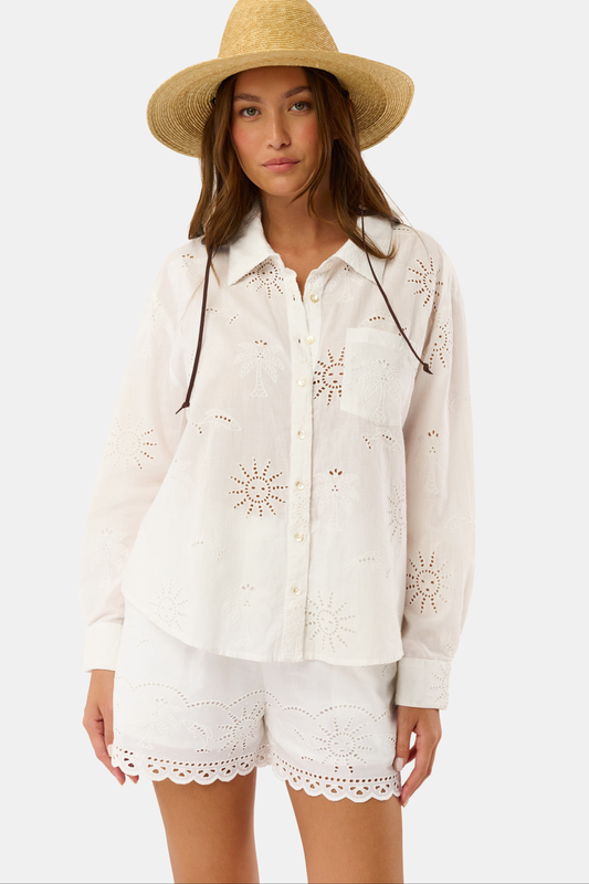 A person wears Roller Rabbit’s Sunny Days Eyelet Cody Shorts—white with an elastic waist, scalloped hems, and intricate cut-out details—and has one hand in the shorts pocket.