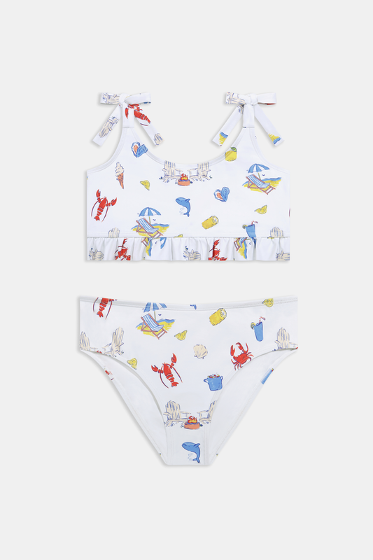 A girl wears the Roller Rabbit Kids Ruffle Catch of the Day Cora Bikini, a white two-piece with a playful print, ruffle trim top, and tie straps, standing against a plain light background.