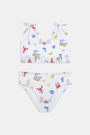 A girl wears the Roller Rabbit Kids Ruffle Catch of the Day Cora Bikini, a white two-piece with a playful print, ruffle trim top, and tie straps, standing against a plain light background.