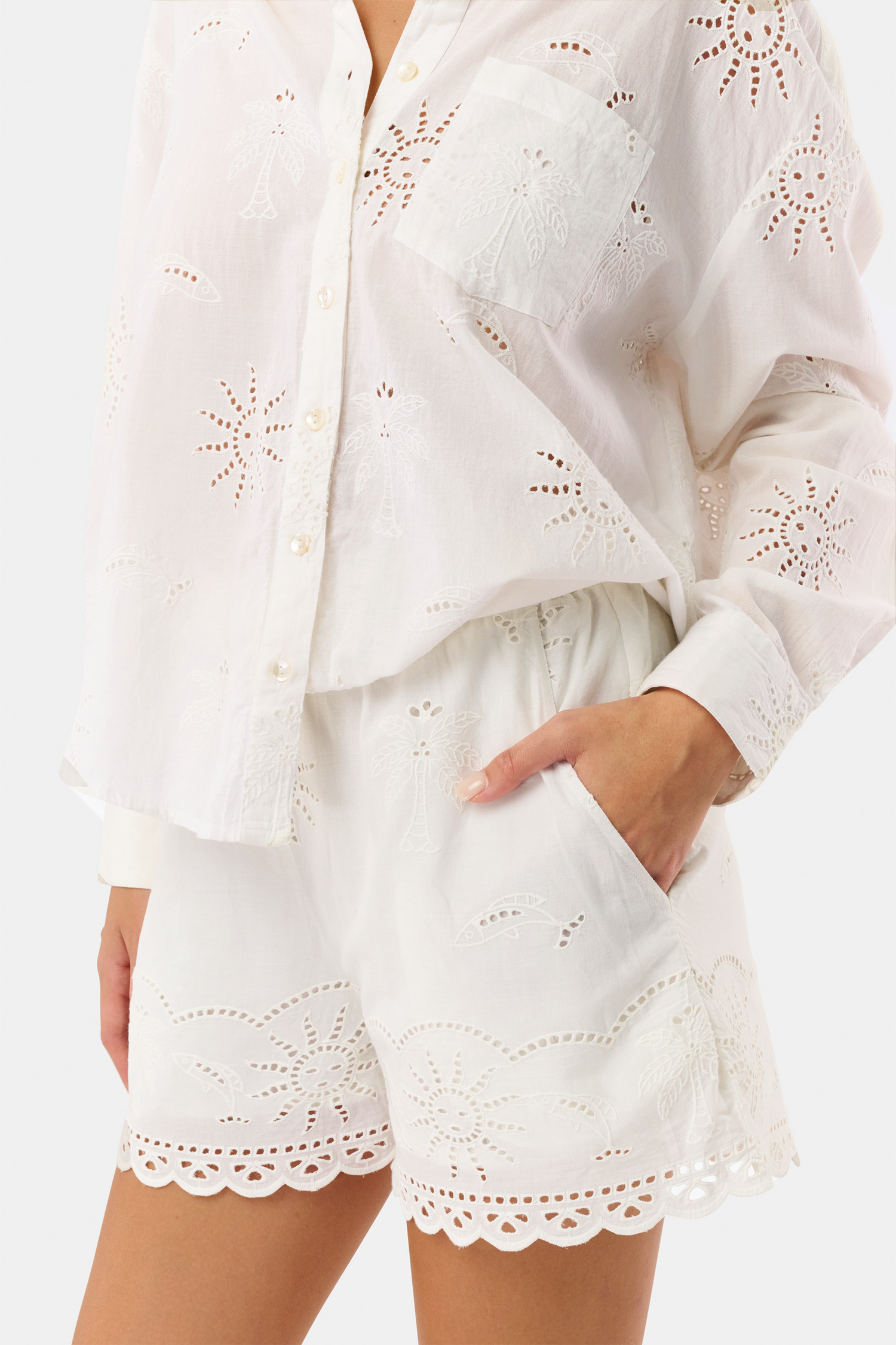 A person wears Roller Rabbit’s Sunny Days Eyelet Cody Shorts—white with an elastic waist, scalloped hems, and intricate cut-out details—and has one hand in the shorts pocket.