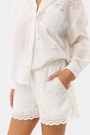 A person wears Roller Rabbit’s Sunny Days Eyelet Cody Shorts—white with an elastic waist, scalloped hems, and intricate cut-out details—and has one hand in the shorts pocket.