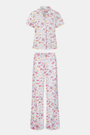 A woman with curly hair models the Roller Rabbit Summer Memories Fiora Pajamas, a Pima cotton set with a colorful sushi print, including a short-sleeve button-up top and long pants, against a white background.
