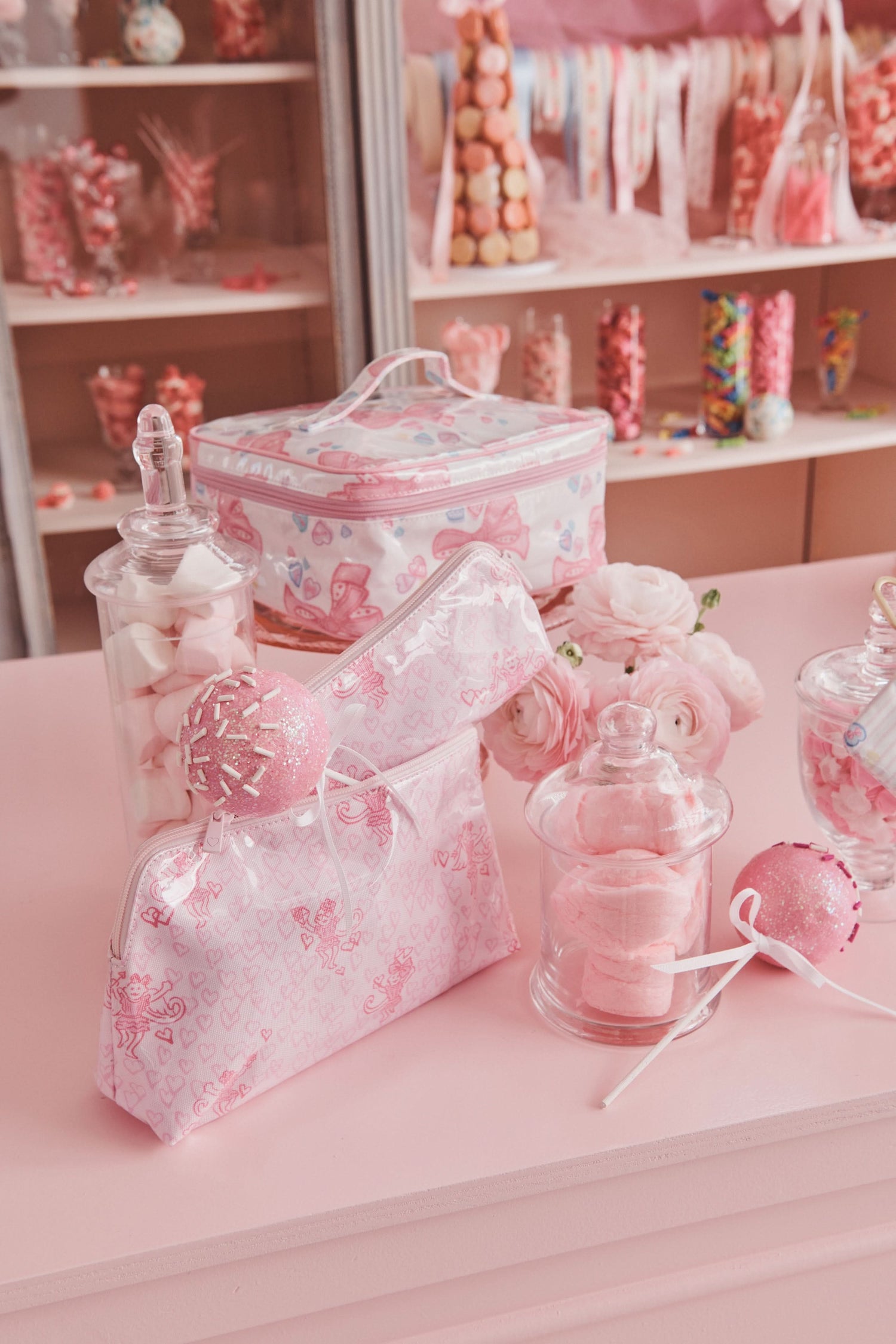 A pastel pink candy shop display features marshmallows, a lollipop, roses, and two Roller Rabbit Cupid's Kiss Makeup Bags with heart patterns on a pink counter, with colorful candy jars in the background.