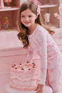 A girl with long brown hair stands against a pale pink wall, wearing Roller Rabbit Kids Cupid's Kiss Juniper Pajamas in light pink and gray socks, looking at the camera with a slight smile.