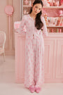 A young woman wearing the Roller Rabbit Cupid's Kiss Boden Pant and fluffy slippers stands in front of a pastel pink, candy-themed counter adorned with jars of sweets and heart decorations.