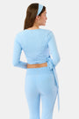 A woman with long brown hair, wearing the Morgan Wrap Top in sky blue with matching patterned leggings and a headband, stands with her back to the camera against a white background, showcasing elegant modern activewear style.