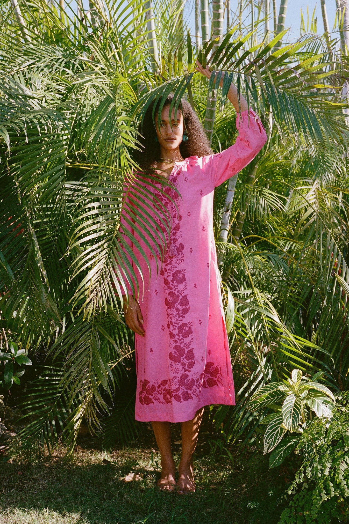A person stands barefoot among lush tropical plants, partly hidden by palm fronds, wearing the bright pink Cross Stitch Vine Shiv Caftan from Roller Rabbit as sunlight filters through the leaves.