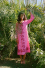 A person stands barefoot among lush tropical plants, partly hidden by palm fronds, wearing the bright pink Cross Stitch Vine Shiv Caftan from Roller Rabbit as sunlight filters through the leaves.