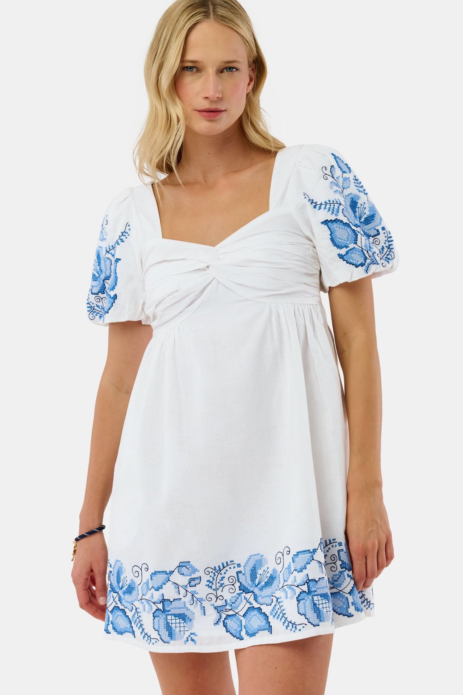 A woman wears the Roller Rabbit Cross Stitch Vine Allora Dress, a short white linen dress with blue floral embroidery on the sleeves and hem, standing with one hand in her pocket against a plain background.