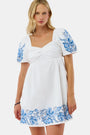 A woman wears the Roller Rabbit Cross Stitch Vine Allora Dress, a short white linen dress with blue floral embroidery on the sleeves and hem, standing with one hand in her pocket against a plain background.