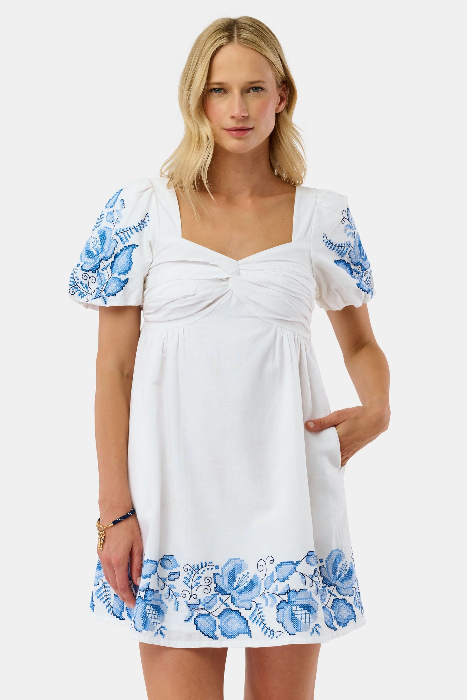A woman wears the Roller Rabbit Cross Stitch Vine Allora Dress, a short white linen dress with blue floral embroidery on the sleeves and hem, standing with one hand in her pocket against a plain background.