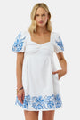 A woman wears the Roller Rabbit Cross Stitch Vine Allora Dress, a short white linen dress with blue floral embroidery on the sleeves and hem, standing with one hand in her pocket against a plain background.