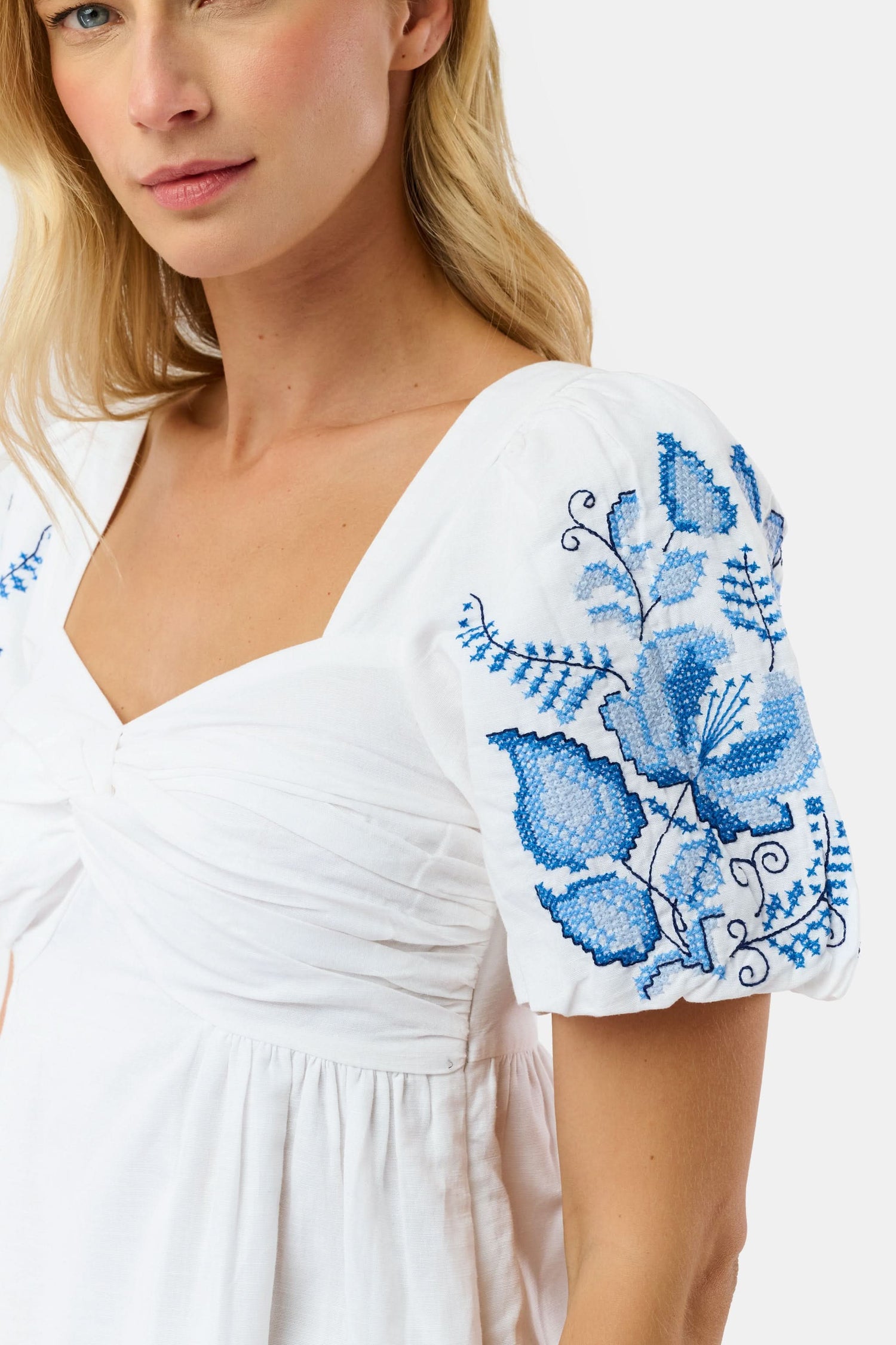 A woman wears the Roller Rabbit Cross Stitch Vine Allora Dress, a short white linen dress with blue floral embroidery on the sleeves and hem, standing with one hand in her pocket against a plain background.