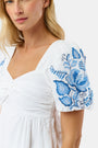 A woman wears the Roller Rabbit Cross Stitch Vine Allora Dress, a short white linen dress with blue floral embroidery on the sleeves and hem, standing with one hand in her pocket against a plain background.
