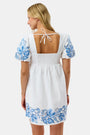 A woman wears the Roller Rabbit Cross Stitch Vine Allora Dress, a short white linen dress with blue floral embroidery on the sleeves and hem, standing with one hand in her pocket against a plain background.
