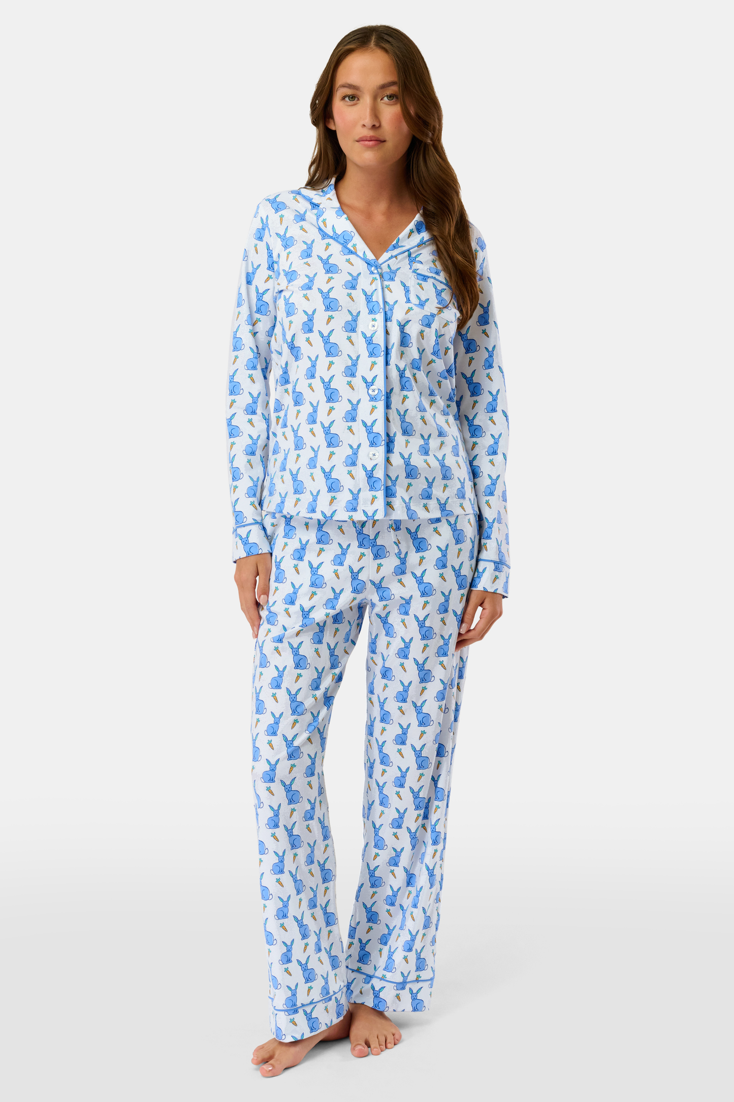A woman with long brown hair stands barefoot in the Roller Rabbit Romy the Rabbit Long Sleeve Polo Pajamas—a matching white set with a blue bunny print, featuring a button-up top and loose-fitting pants. She has a neutral expression.