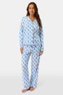 A woman with long brown hair stands barefoot in the Roller Rabbit Romy the Rabbit Long Sleeve Polo Pajamas—a matching white set with a blue bunny print, featuring a button-up top and loose-fitting pants. She has a neutral expression.