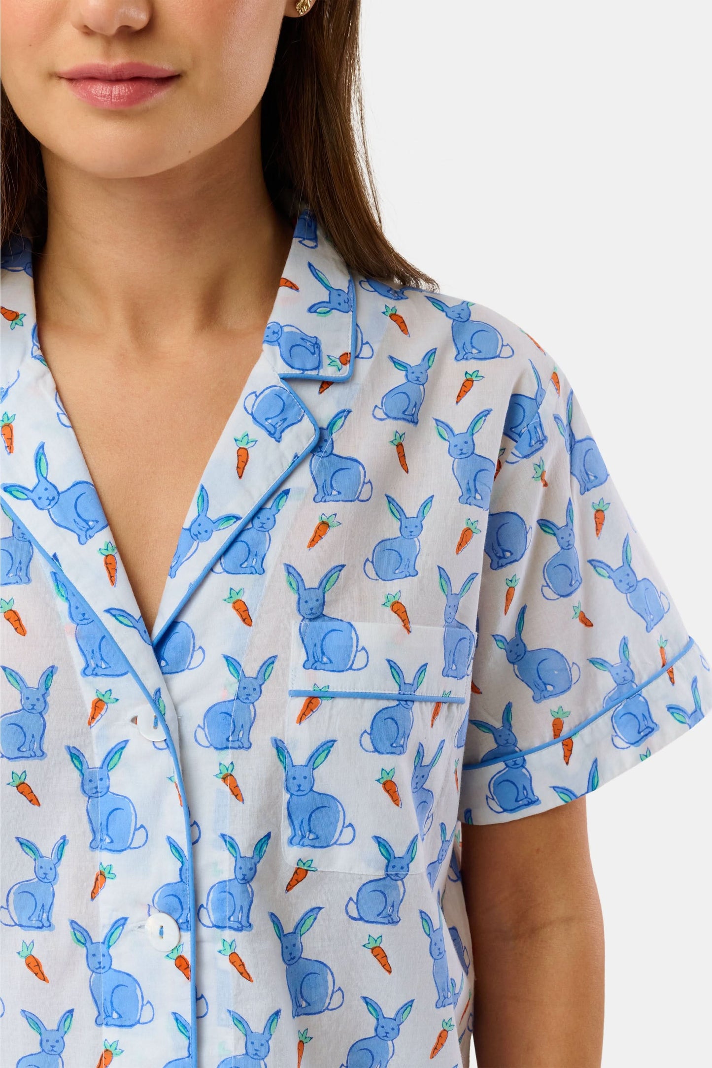 A woman wears the Roller Rabbit Romy the Rabbit Livy Set, a light blue cotton two-piece loungewear featuring a colorful bunny print, as she stands smiling against a plain white background.