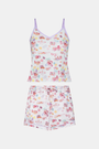 A woman wears the Roller Rabbit Summer Memories Mischa Set, a 100 Pima cotton cami tank and boxer set with a pastel travel icon print on white, standing against a light background.