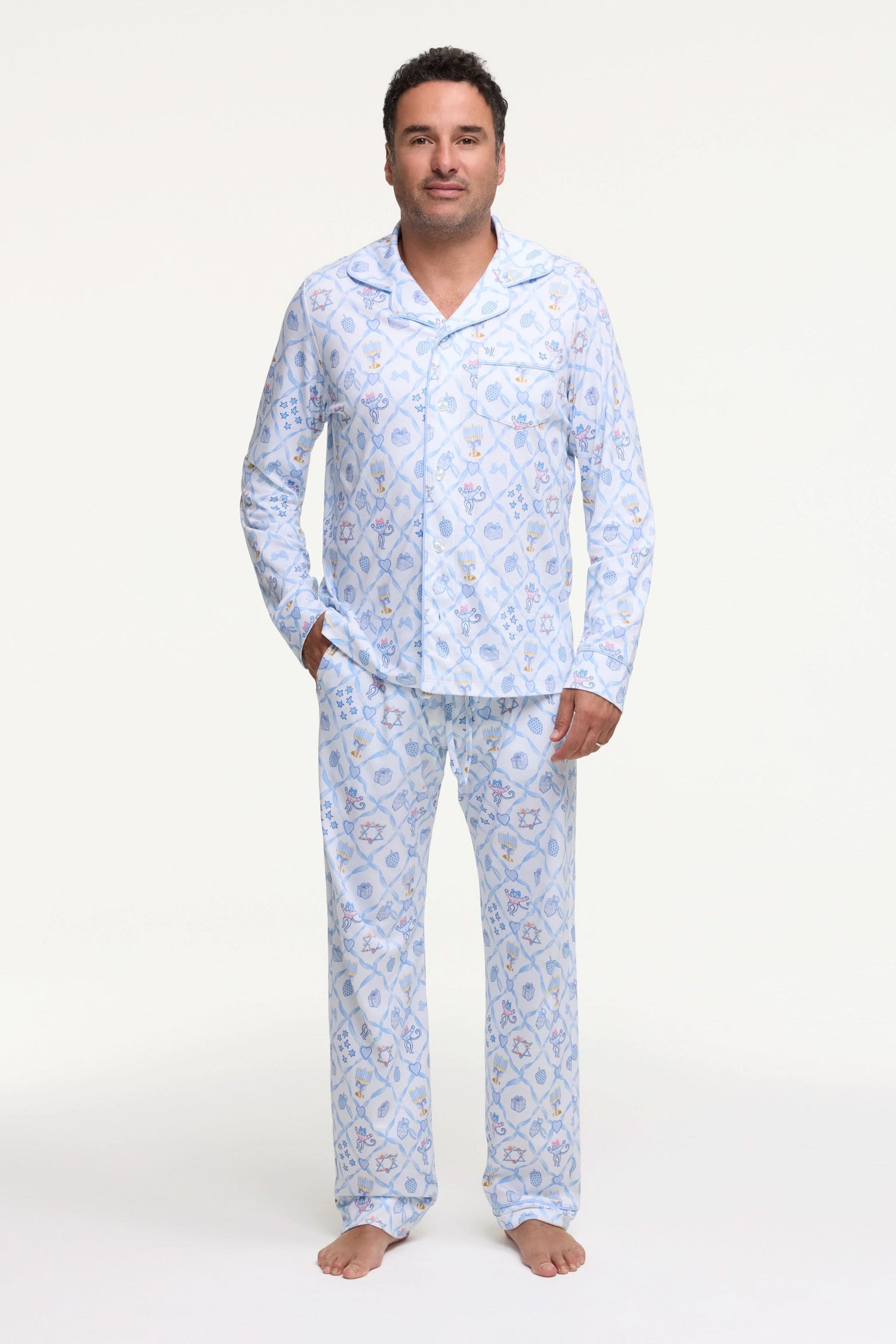 A man stands barefoot on a white background wearing Roller Rabbit's Mens Starry Nights Lattice Long Sleeve Polo Pajamas, featuring light blue and white patterns. He smiles with one hand in his pocket, highlighting the comfort of these matching PJs.