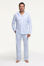 A man stands barefoot on a white background wearing Roller Rabbit's Mens Starry Nights Lattice Long Sleeve Polo Pajamas, featuring light blue and white patterns. He smiles with one hand in his pocket, highlighting the comfort of these matching PJs.