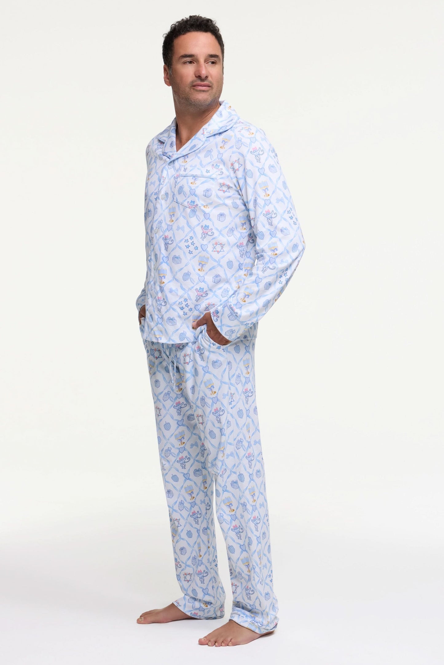 A man stands barefoot on a white background wearing Roller Rabbit's Mens Starry Nights Lattice Long Sleeve Polo Pajamas, featuring light blue and white patterns. He smiles with one hand in his pocket, highlighting the comfort of these matching PJs.